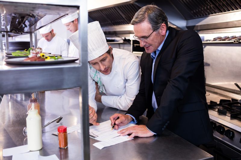 male-restaurant-manager-writing-clipboard-while-interacting-head-chef